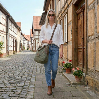 Blonde Frau im Urban-Look geht auf einer historischen kopfsteinpflaster Straße und trägt eine olivgrüne Canvas Crossbody-Bag am langen Riemen über der Schulter – Großer Damen-Shopper aus Segeltuch, kombiniert mit blauer Jeans und weißer Bluse.