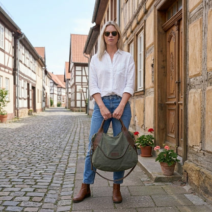 Blonde Frau im Urban-Look steht frontal auf einer historischen kopfsteinpflaster Straße und hält eine Armeegrün Canvas Umhängetasche Vintage mit beiden Händen – Großer Damen-Shopper aus Segeltuch, kombiniert mit blauer Jeans und weißer Bluse