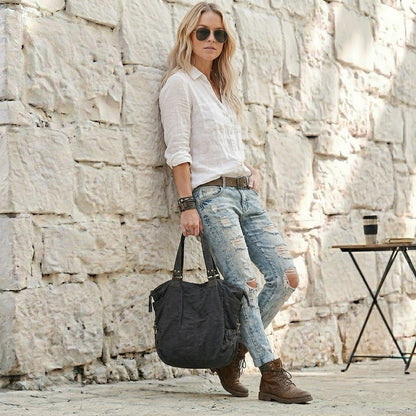 Frau im Casual-Look lehnt an einer weißen Steinwand und hält eine schwarze Canvas Damentasche in der Hand – Großer Damen-Shopper aus Segeltuch, kombiniert mit destroyed Jeans und weißer Bluse.