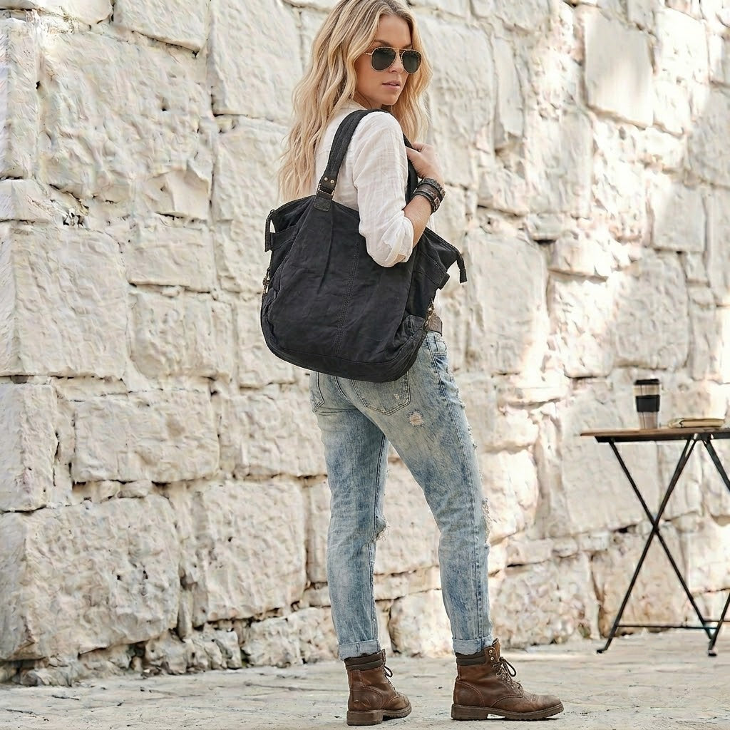 Blonde Frau im Urban-Look steht im Halbdrehung vor einer weißen Steinwand und trägt eine schwarze Canvas Umhängetasche über der Schulter – Großer Damen-Shopper aus Segeltuch, kombiniert mit destroyed Jeans und weißer Bluse