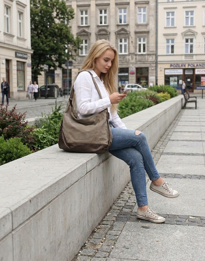 Moderne Frau sitzt mit der khakibraunen Hobo Bag aus Segeltuch auf einer Steinstufe in der Stadt und nutzt ihr Smartphone im Freizeitlook.