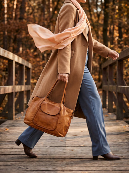 Eine Frau in einem braunen Mantel geht durch den Park und hält eine cognacbraune echtleder shopper -schultertasche in der Hand