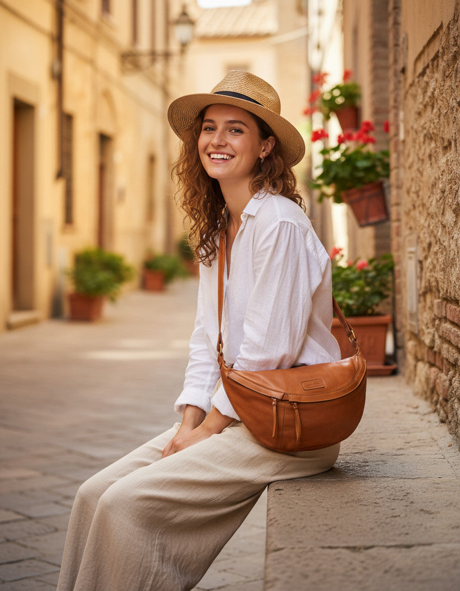 Urlaubs-Inspiration aus Italien: Frau sitzt in weißer Leinenbluse auf alten Steinstufen mit der cognacfarbenen XXL Crossbody-Tasche aus Kalbsleder