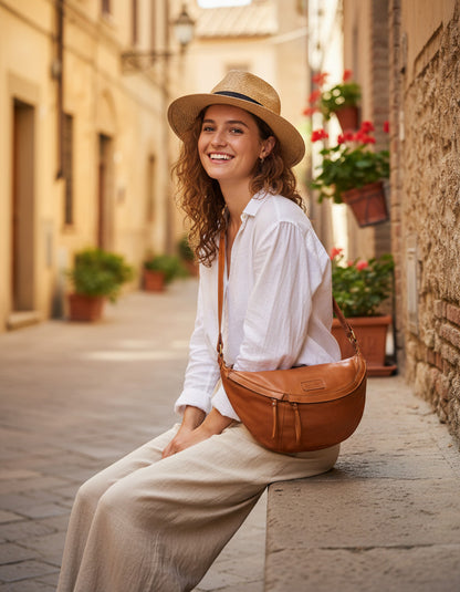 Urlaubs-Inspiration aus Italien: Frau sitzt in weißer Leinenbluse auf alten Steinstufen mit der cognacfarbenen XXL Crossbody-Tasche aus Kalbsleder